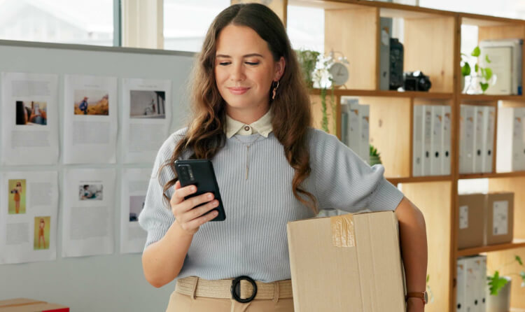 woman smiling business owner looking at phone holding box
